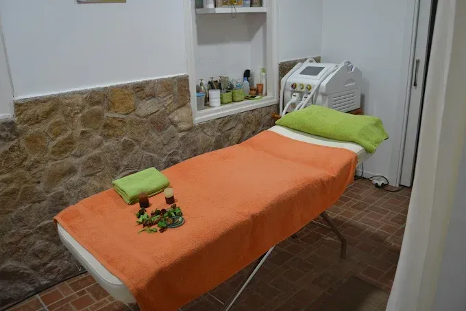 Sala de spa con camilla de masaje con tapizado naranja, almohada y toalla verdes, y detalle de pared de piedra. Estantes con productos y una máquina blanca en la esquina.
