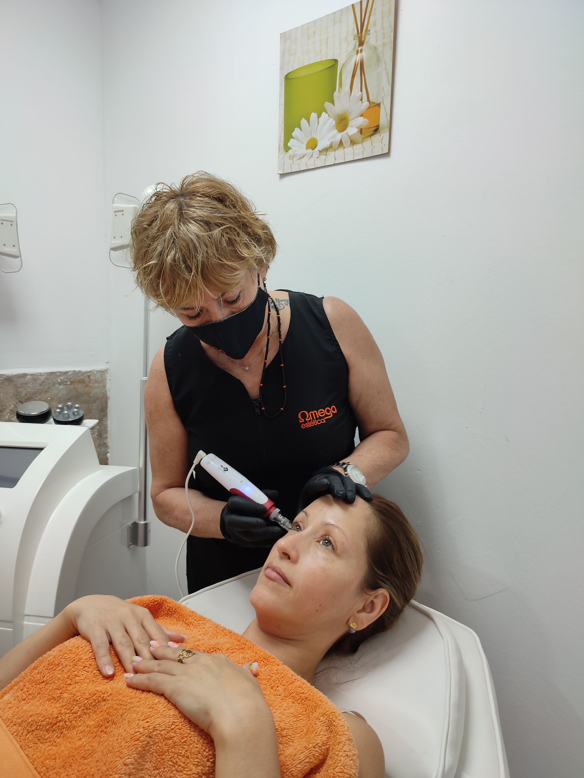 Mujer recibiendo un tratamiento de cejas en un spa. El técnico usa mascarilla y guantes, sosteniendo un instrumento similar a un bolígrafo cerca de la frente de la clienta.