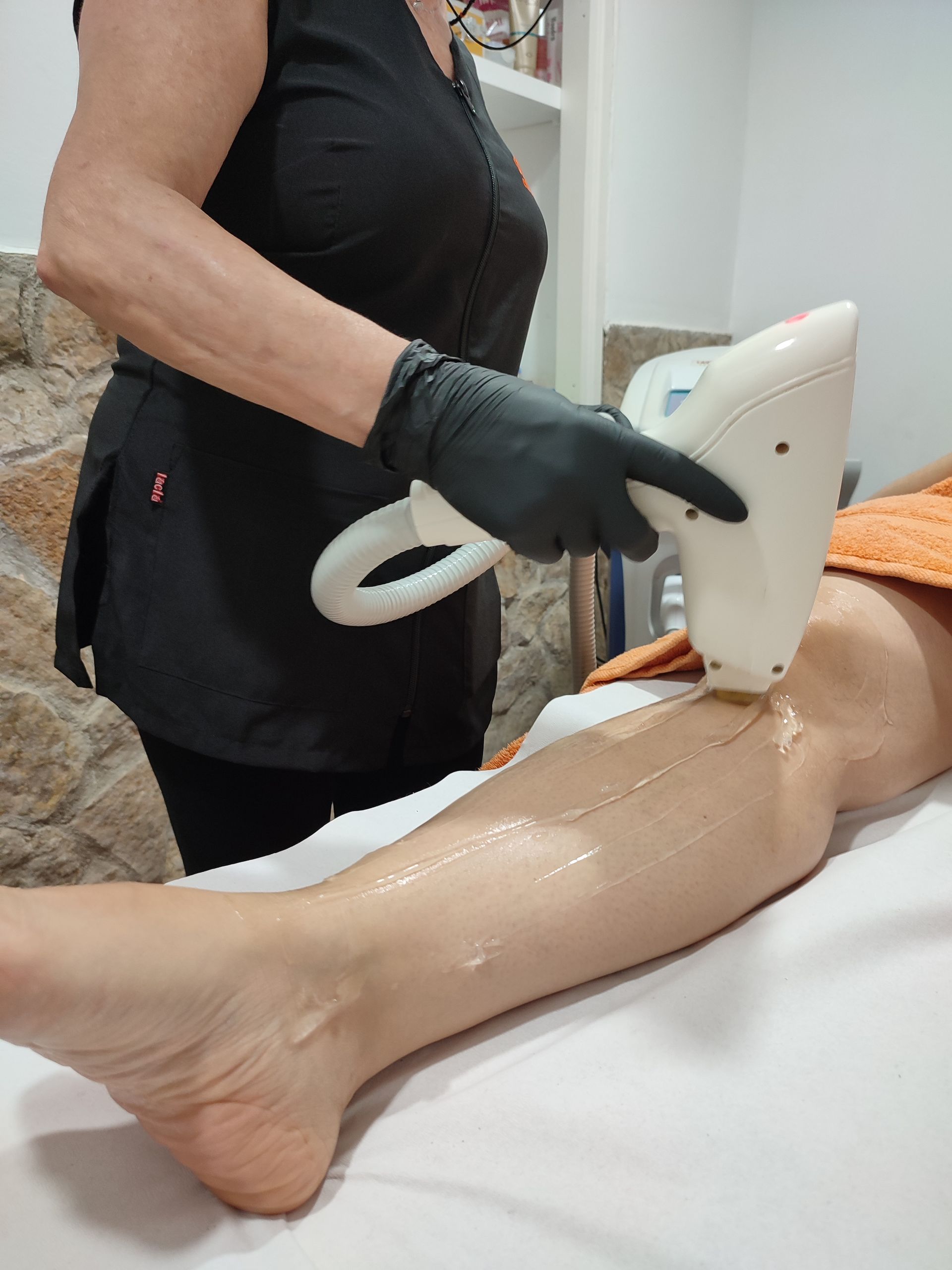 Persona que realiza depilación láser en una pierna en un spa. El técnico lleva guantes negros y sostiene un dispositivo blanco.