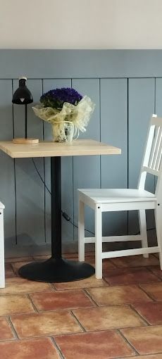 Una pequeña mesa cuadrada con flores y una silla blanca contra una pared revestida de paneles azules. El suelo es de baldosas rojas.