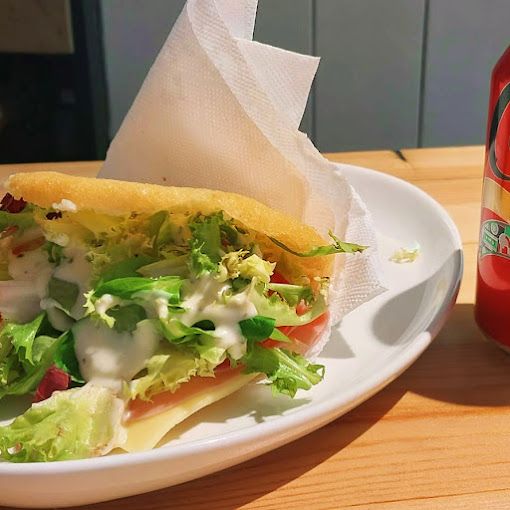 Sándwich con lechuga, tomate y salsa blanca en un plato blanco, con una bebida de fondo, todo sobre una mesa de madera.