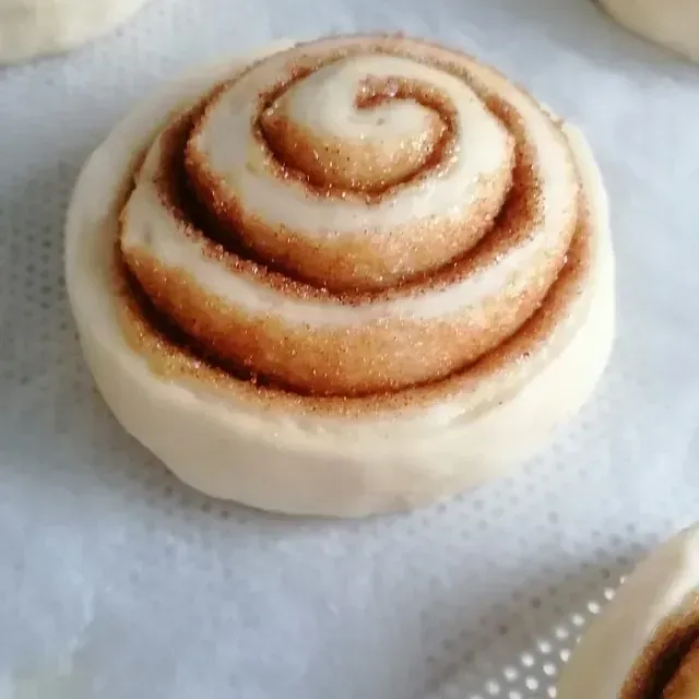 Primer plano de un rollo de canela relleno en espiral, sobre una superficie blanca. El rollo es pálido y presenta un remolino de canela y azúcar.