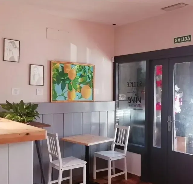 Interior de una cafetería con dos sillas blancas en una mesa pequeña. Un cuadro de limones cuelga en la pared y un cartel de 