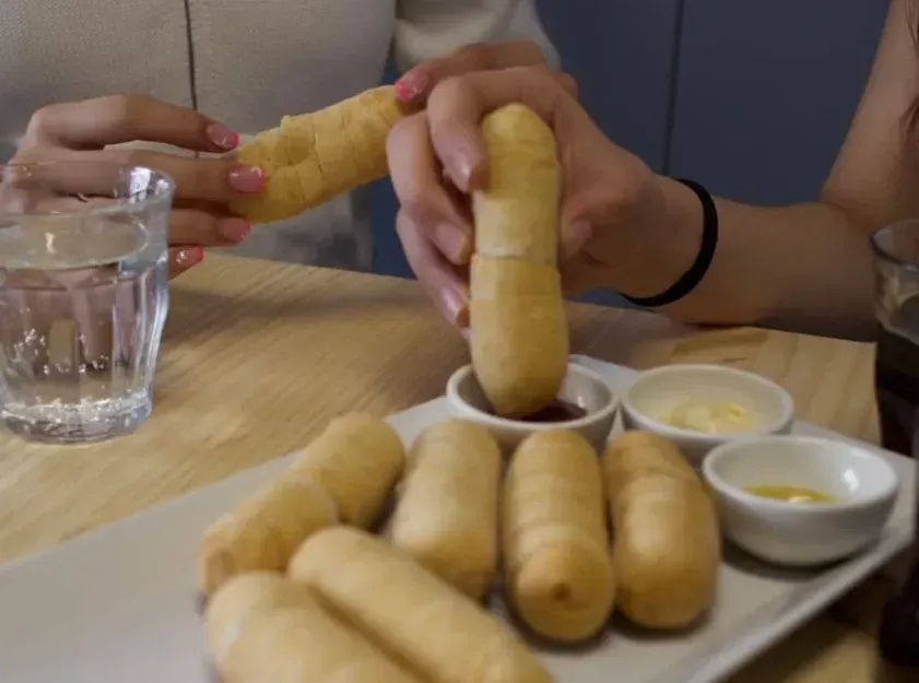 Persona mojando un tequeño (un palito de queso frito) en un plato pequeño con salsa. La comida se sirve en un plato con otros tequeños y salsas para mojar.