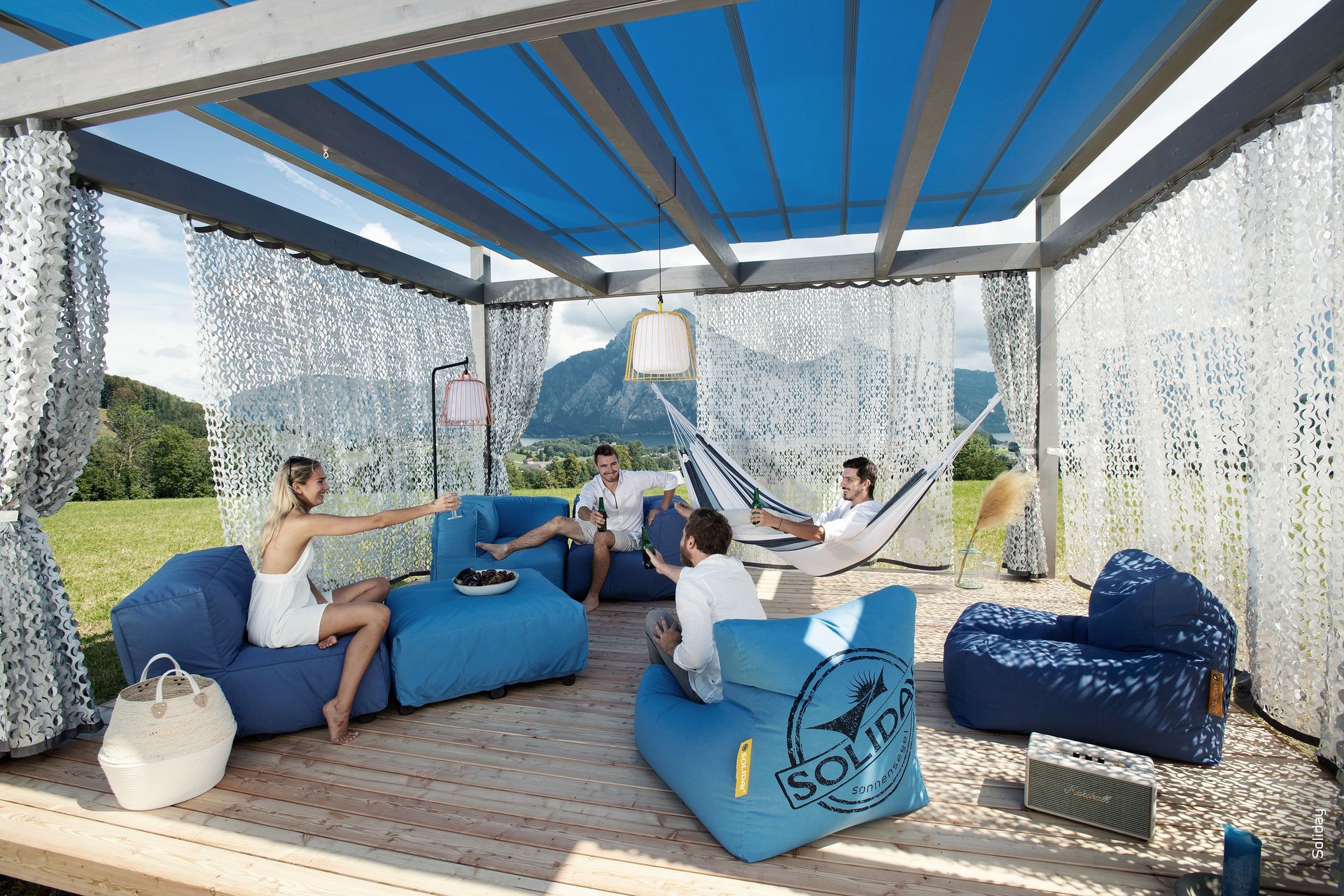 Modernes zweistöckiges Haus mit heller Holzverkleidung, großen Fenstern und Sitzgelegenheiten im Freien unter einem Sonnensegel.