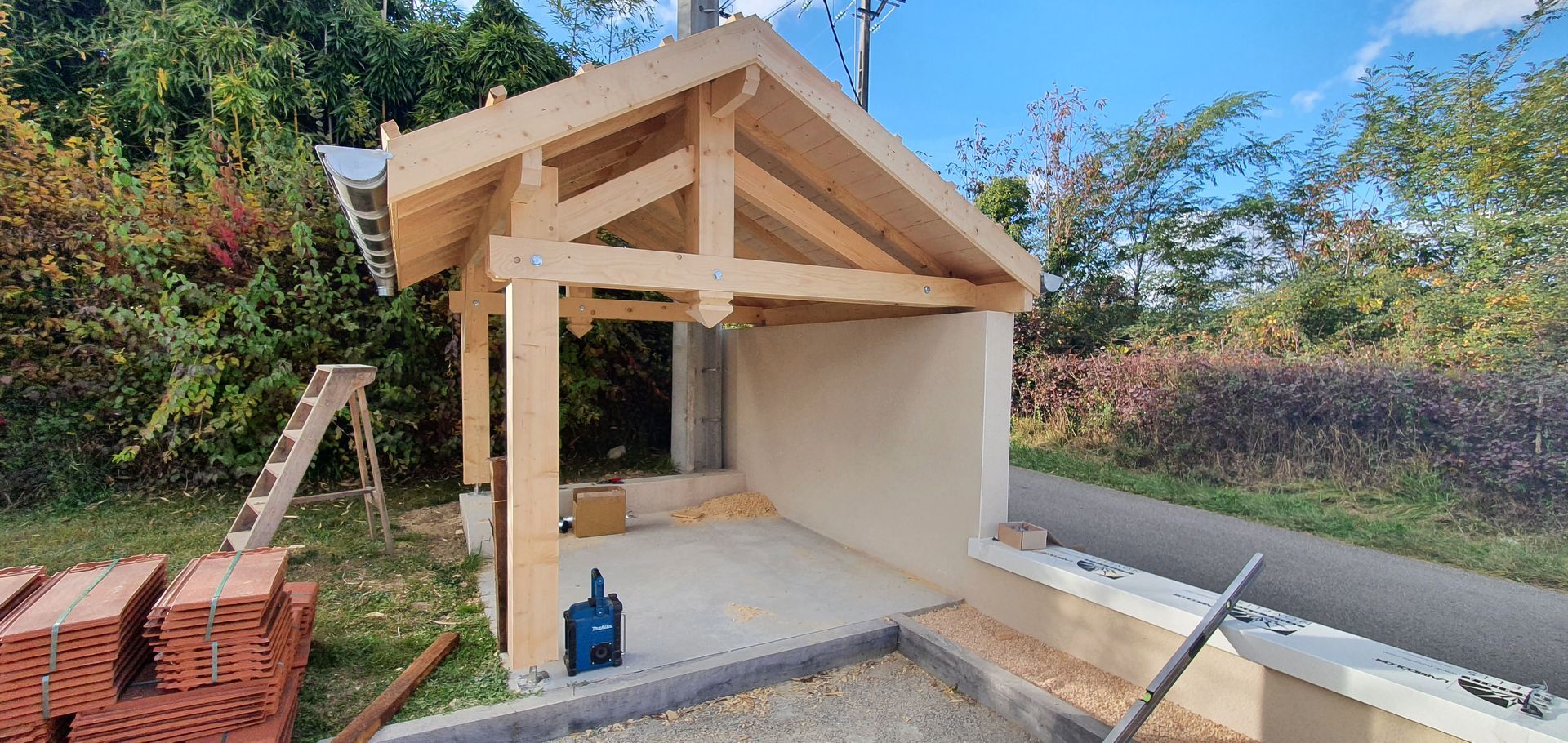 Structure en bois en construction, avec un plancher en béton, entourée de verdure et d'une route.