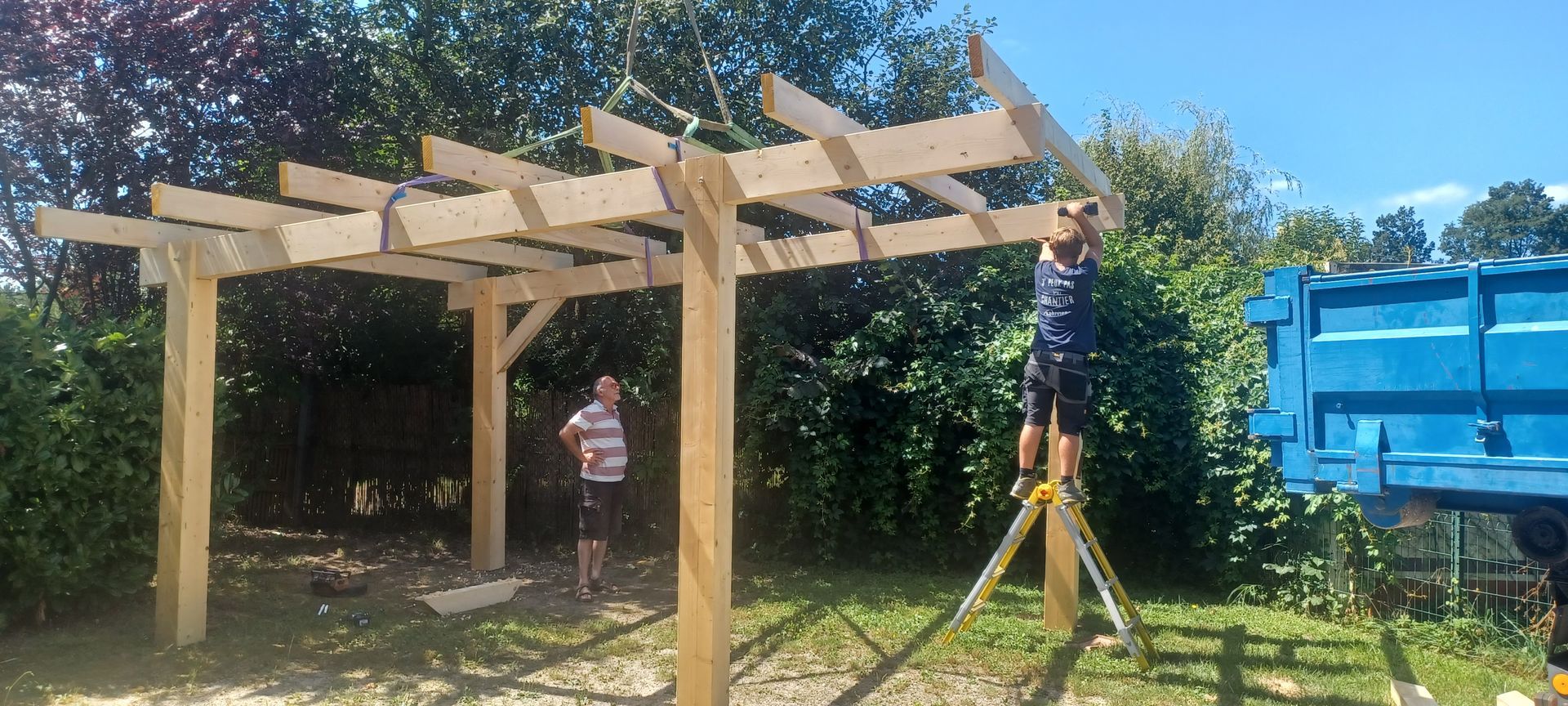 Des gens construisent une pergola en bois dans une cour ; une personne sur une échelle.