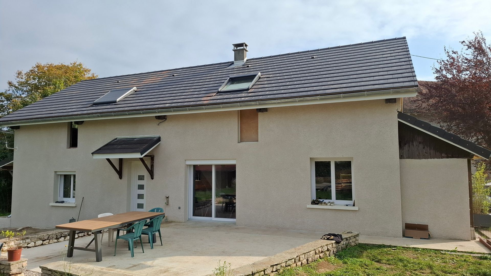 Maison à deux étages avec toit gris, murs clairs et terrasse avec table et chaises. Ciel nuageux.