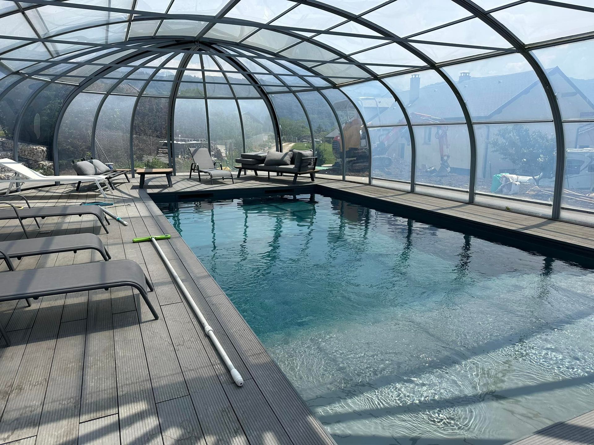 Piscine couverte avec toit arqué transparent ; terrasse grise et chaises longues entourent l'eau bleue.