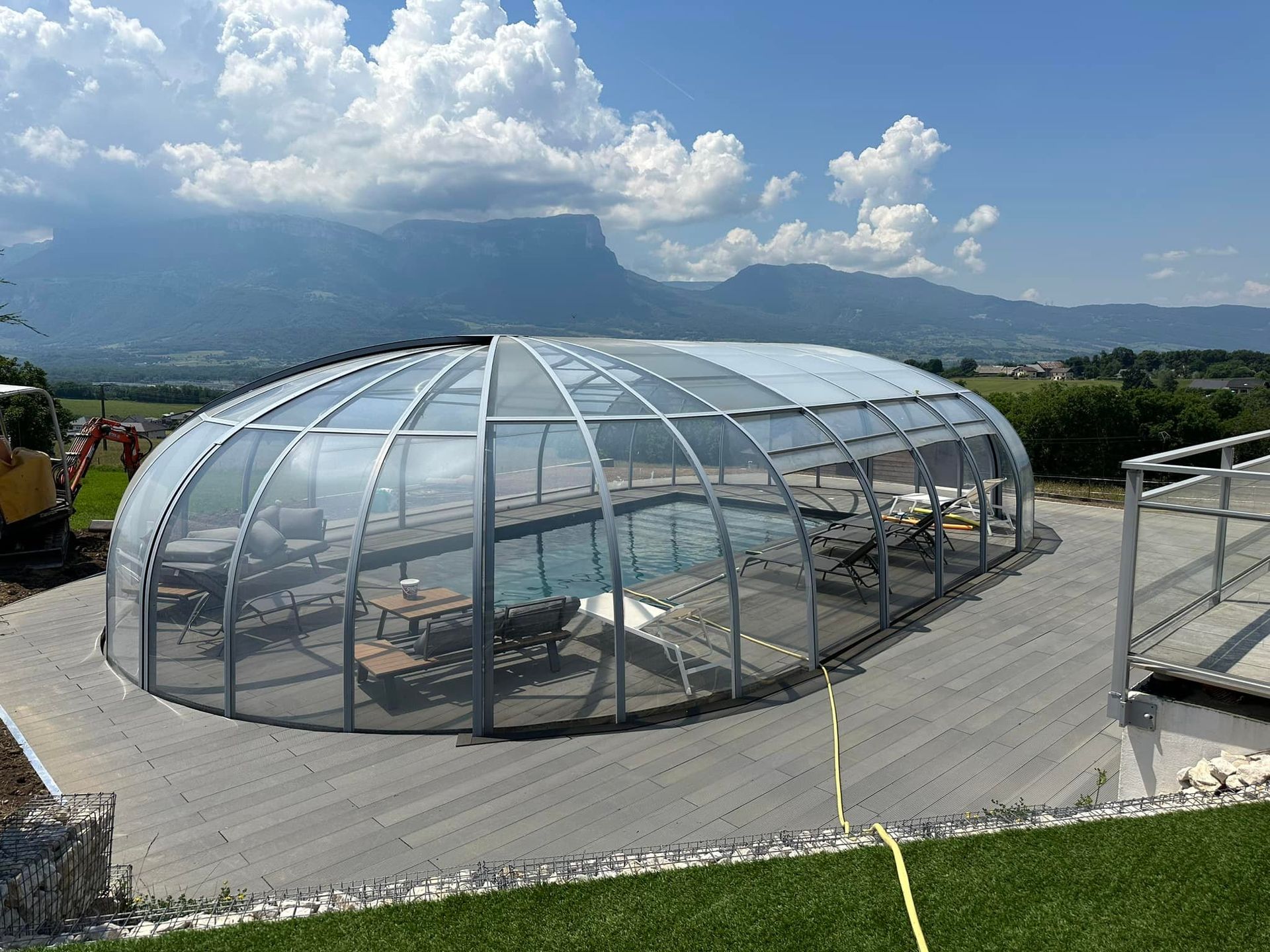 Piscine couverte par une structure en forme de dôme transparent avec tables et chaises, montagnes en arrière-plan.
