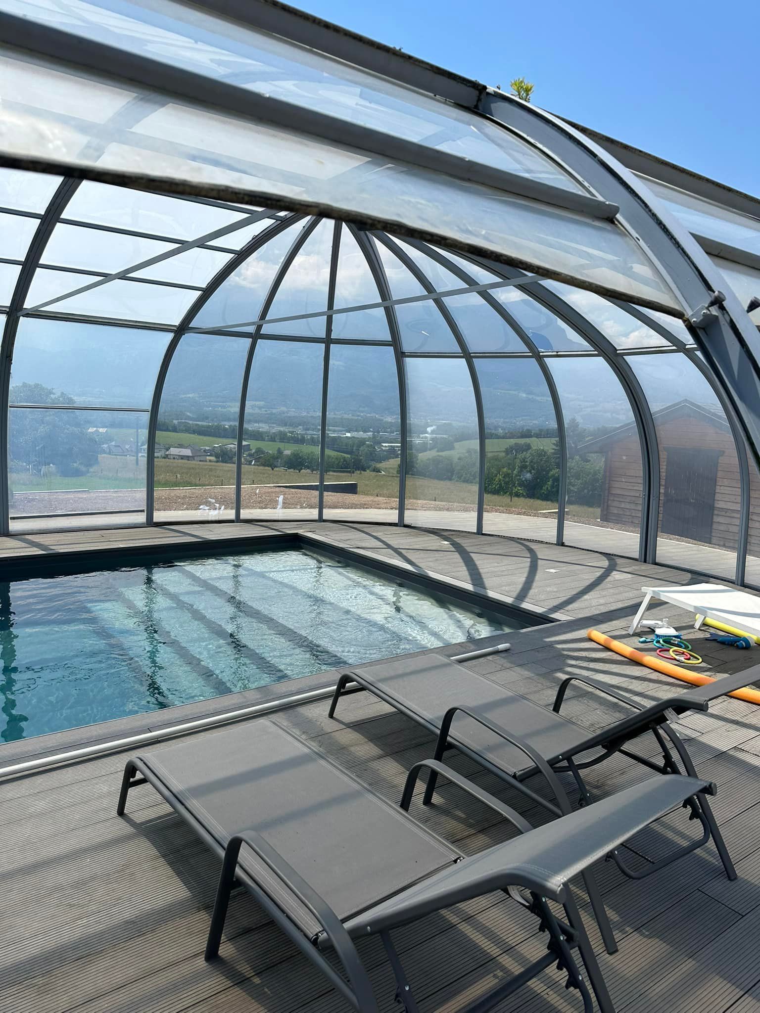 Piscine avec couverture rétractable, deux chaises longues sur une terrasse en bois, vue sur les montagnes.
