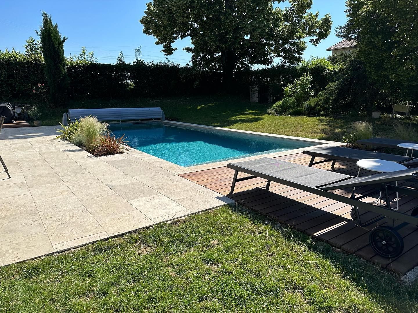 Piscine à l'eau bleue, entourée de pierre beige et d'herbe verte, avec chaises longues.