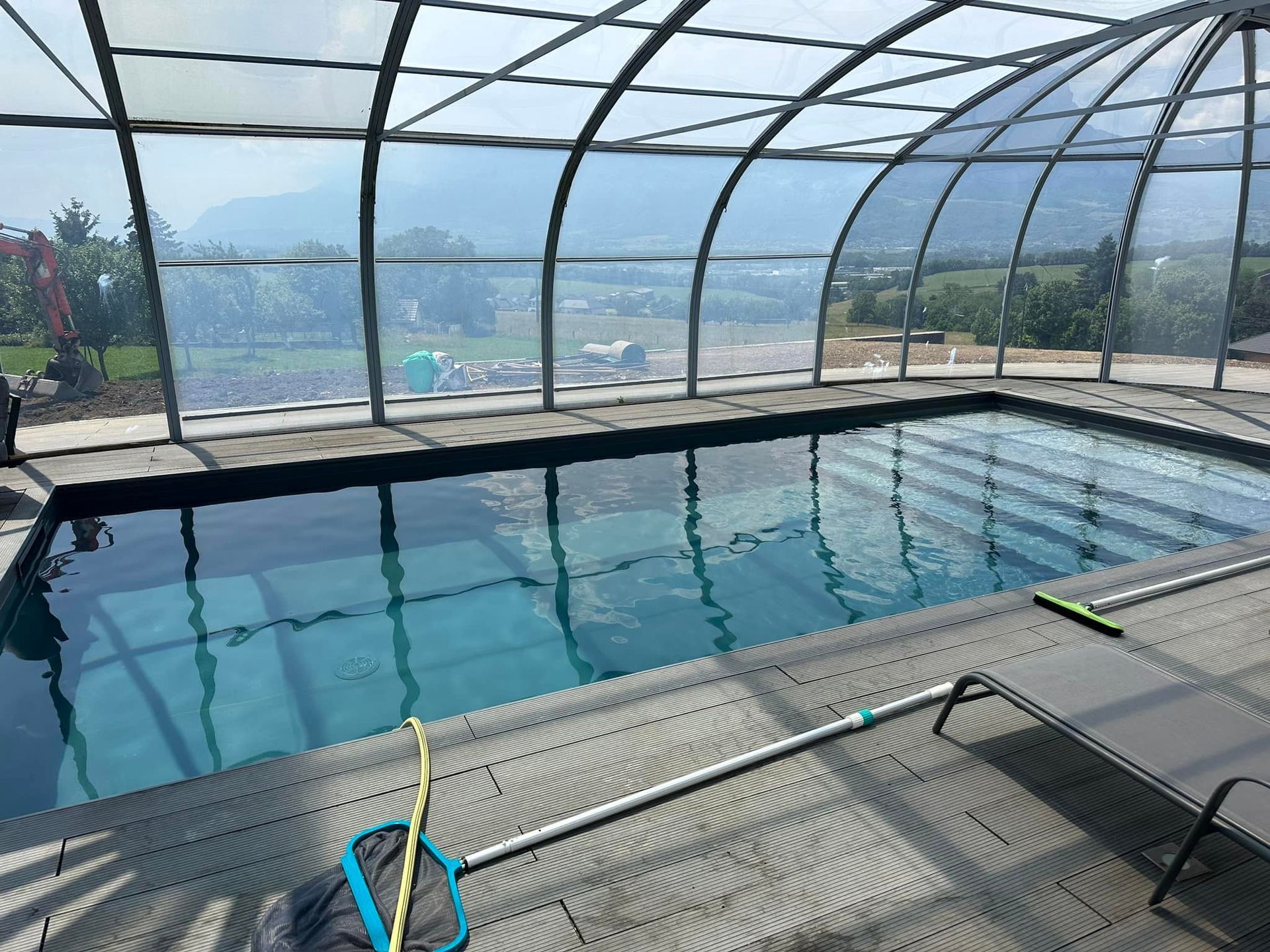 Piscine couverte par une verrière avec des outils de nettoyage au bord, vue sur paysage.