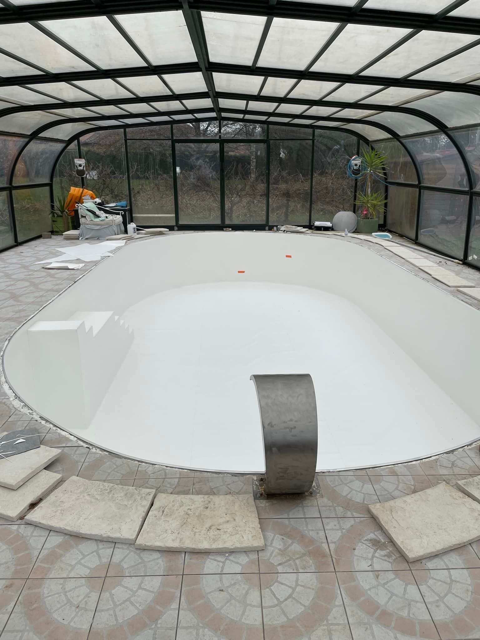 Piscine couverte en construction, murs blancs, sol en béton, toiture métallique noire.