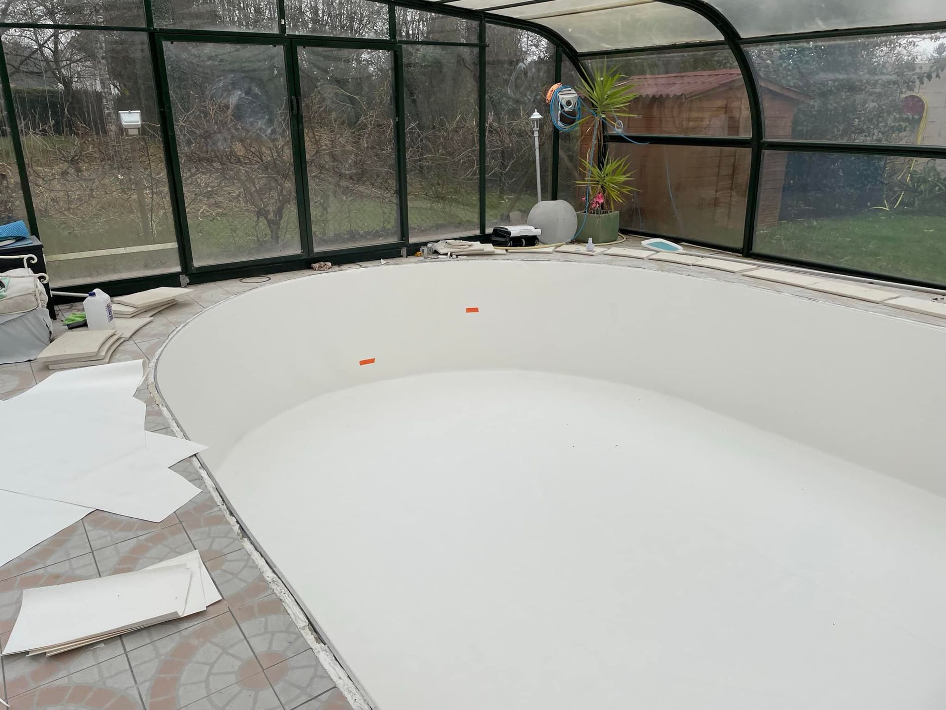 Piscine couverte en construction, murs blancs, sol en béton, toiture métallique noire.