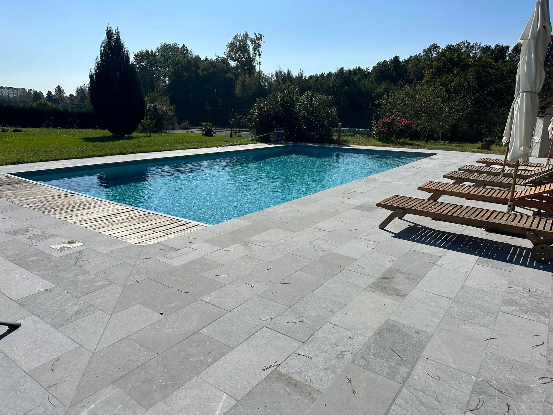 Piscine avec eau bleue, terrasse en pierre et chaises longues par une journée ensoleillée dans un cadre d'arrière-cour.