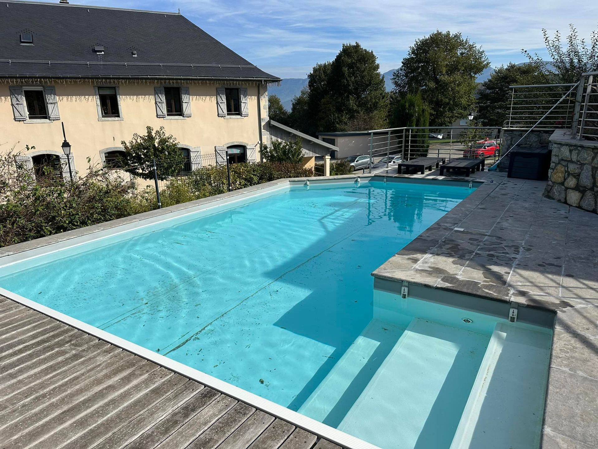 Une piscine rectangulaire avec marches, terrasse en bois, eau claire, maison en pierre en arrière-plan par une journée ensoleillée.