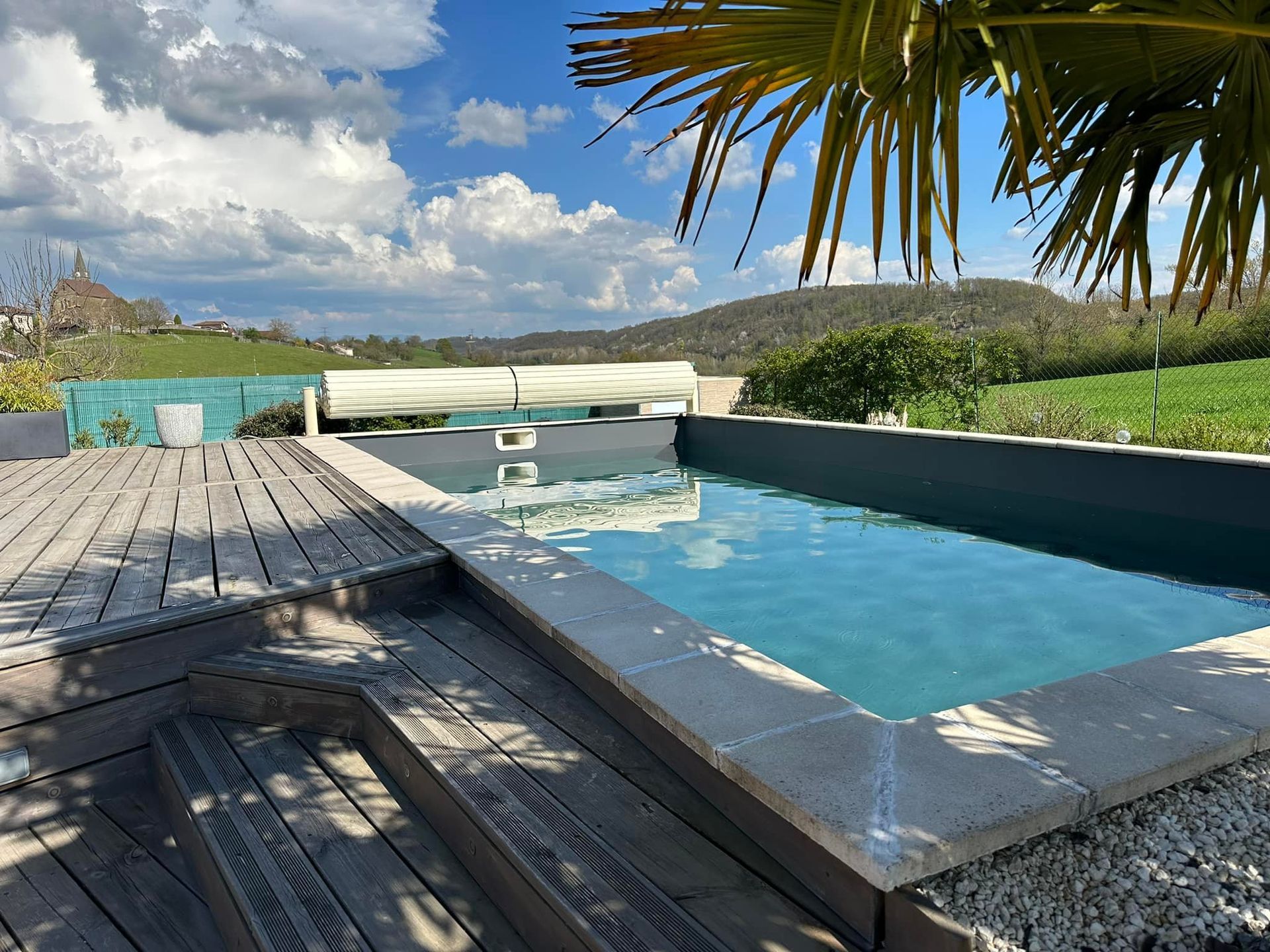 Piscine avec terrasse en bois, journée ensoleillée avec vue sur la montagne, palmier, eau bleue.