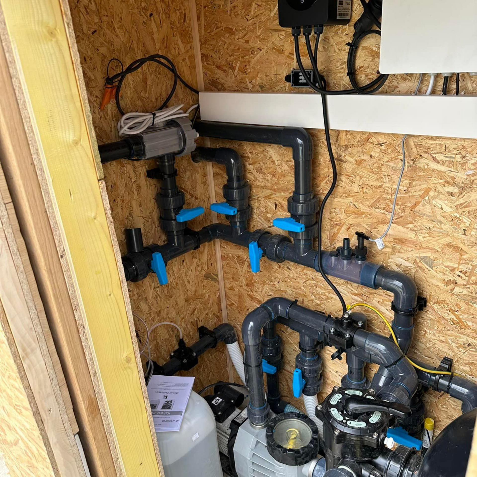 Système de plomberie avec poignées de vannes bleues et tuyaux noirs à l'intérieur d'un local en bois.