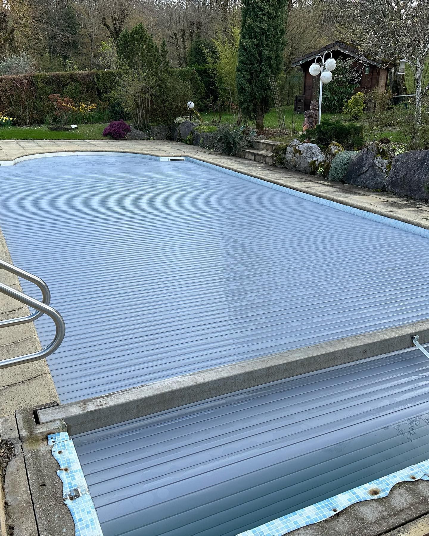 Piscine couverte d'une protection bleue, entourée de pierres, d'arbres et d'un jardin.