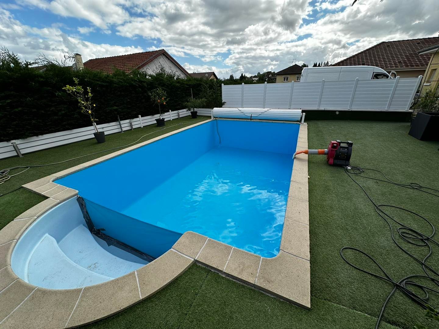 Piscine rectangulaire bleue avec marches, entourée de pelouse verte et d'une clôture blanche, sous un ciel partiellement nuageux.