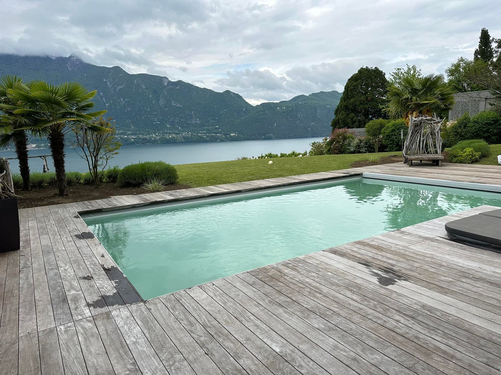 Vue sur la piscine, un lac et des montagnes ; une terrasse en bois et des palmiers encadrent l'eau turquoise.
