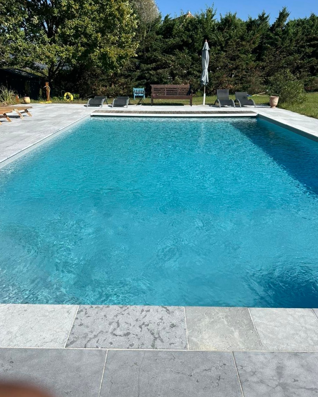 Piscine à l'eau bleu clair, entourée de pierre grise et de verdure.
