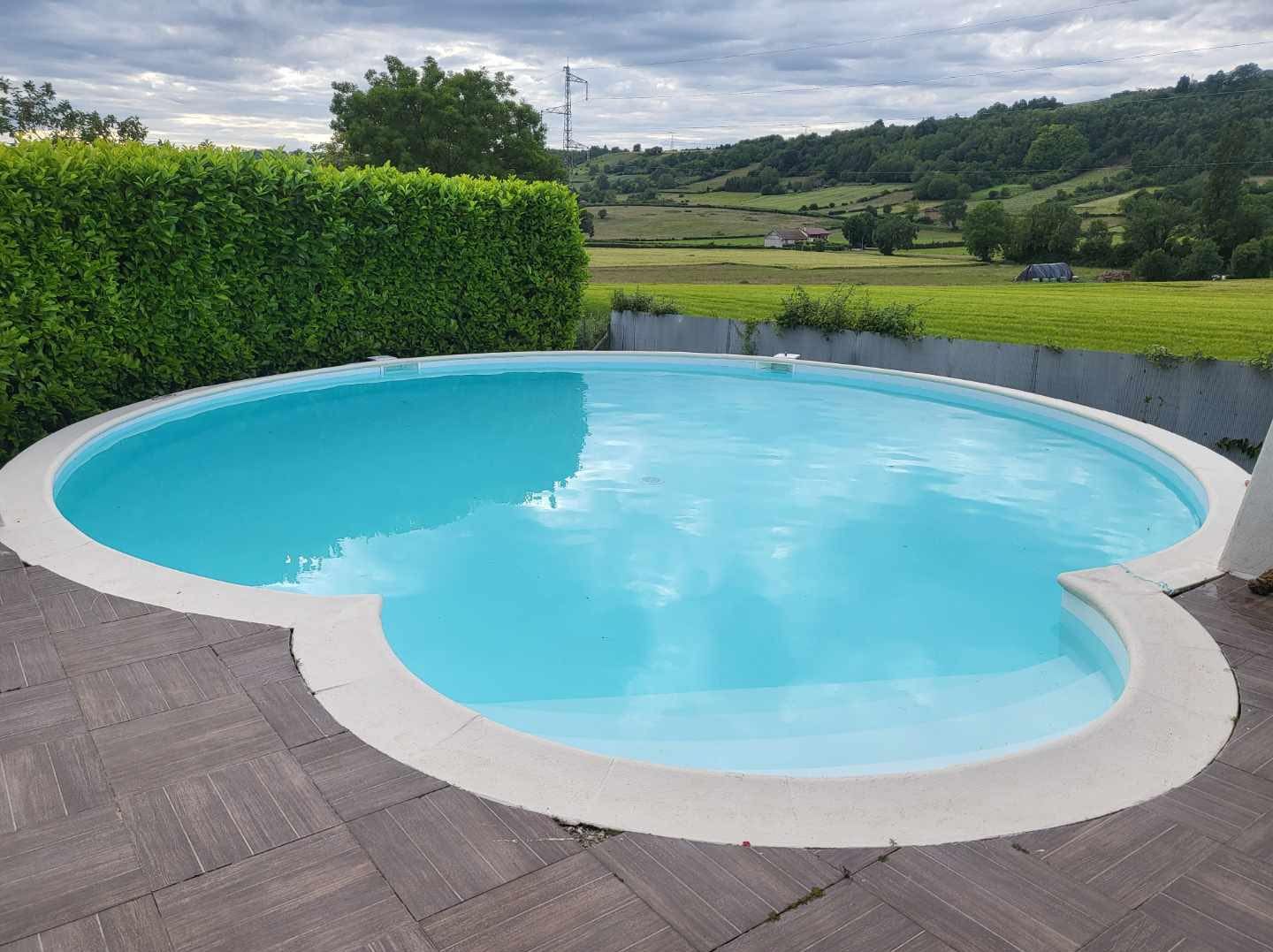 Piscine ovale à l'eau bleu clair, margelle en pierre, dans une cour arrière de maison.