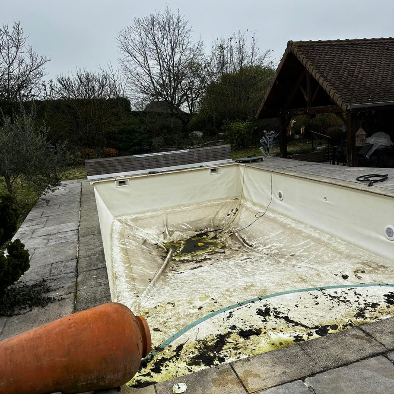 Une piscine vide et sale avec des débris, entourée de pierres et d'un pavillon extérieur par une journée nuageuse.