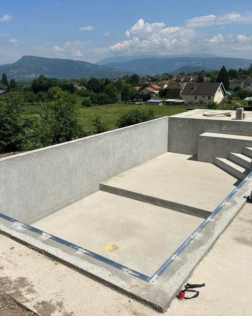 Piscine en béton en construction avec marches, donnant sur un paysage verdoyant et des maisons sous un ciel bleu.