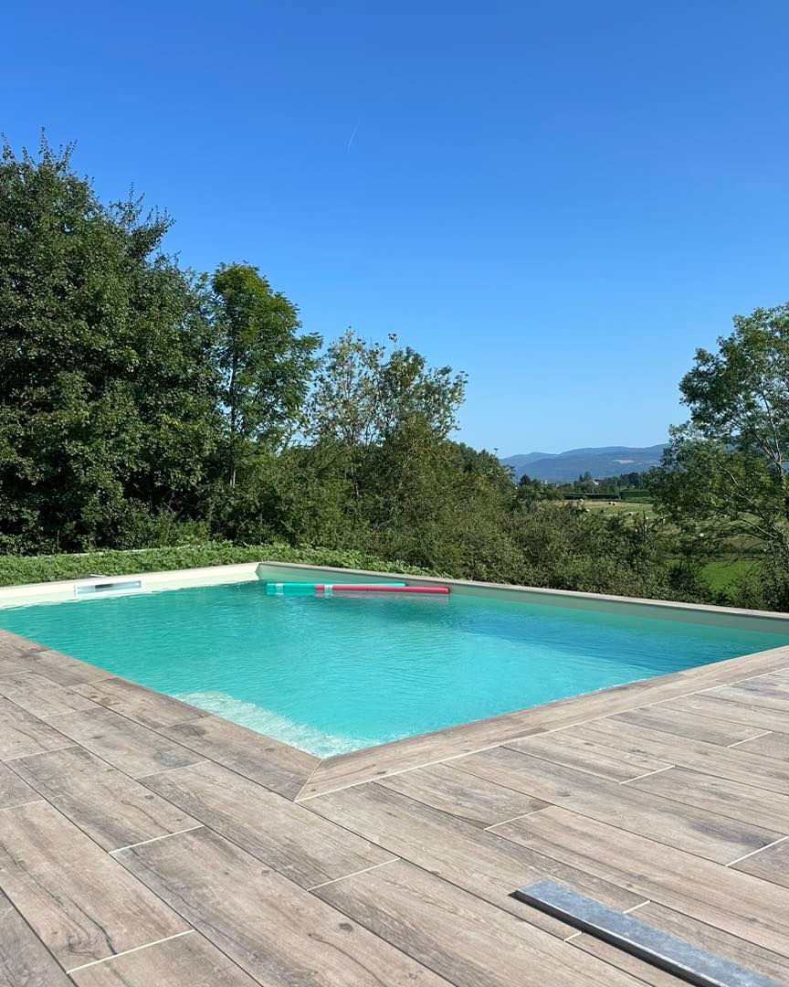 Piscine avec eau turquoise et terrasse en bois, entourée d'arbres sous un ciel bleu clair.
