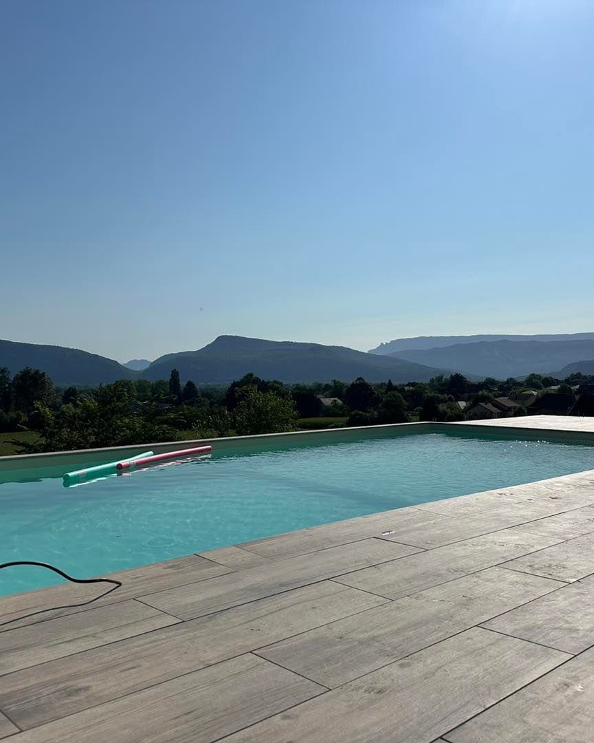 Piscine à débordement avec vue sur les montagnes par une journée ensoleillée. Ciel bleu et eau turquoise. Terrasse en bois.