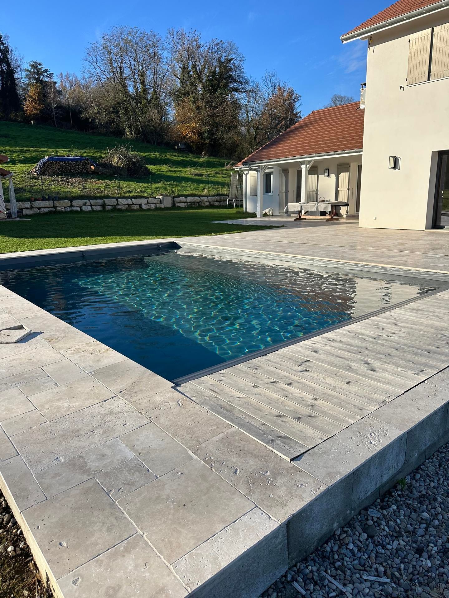 Une piscine rectangulaire avec un intérieur bleu foncé entouré de carreaux de pierre grise, à côté d'une maison par une journée ensoleillée.