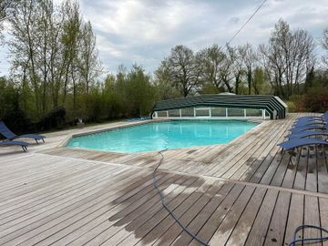 Piscine avec terrasse en bois, recouverte d'un auvent vert et blanc, entourée d'arbres.