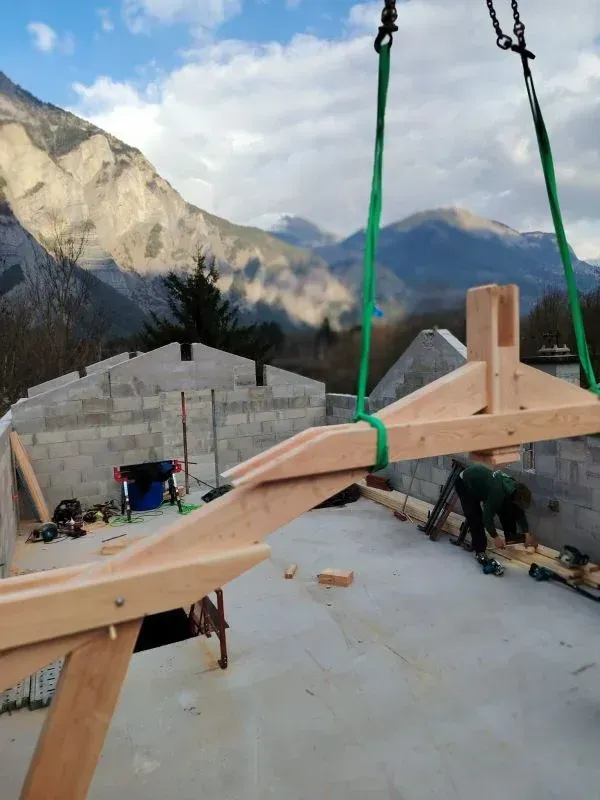 Une poutre de toit en bois est soulevée par des sangles vertes sur un chantier. Les montagnes en arrière-plan.