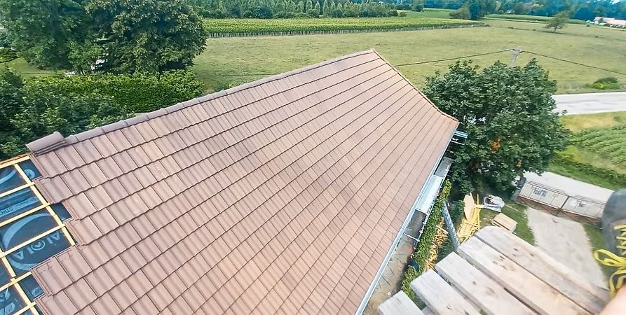 Toiture en tuiles brunes en construction, arbres et champs verts en arrière-plan.