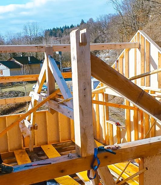 Maison en bois en construction, charpente visible, avec soleil et ciel bleu.
