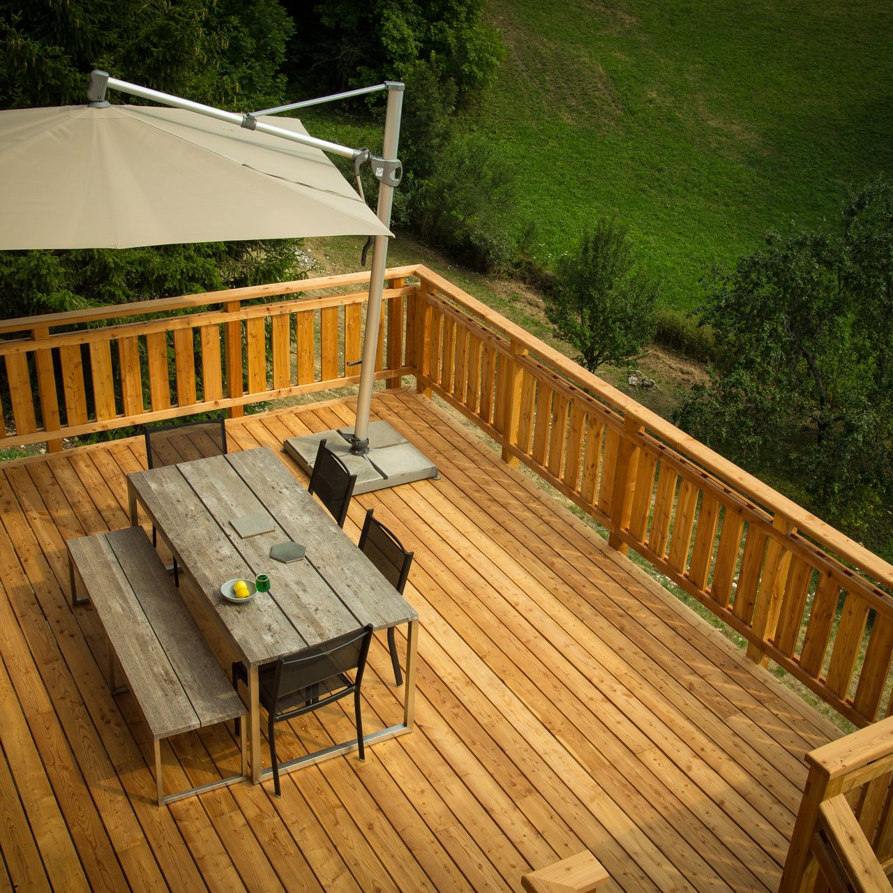 Terrasse en bois surélevée avec table, bancs, chaises et parasol. Vue sur une colline verdoyante.