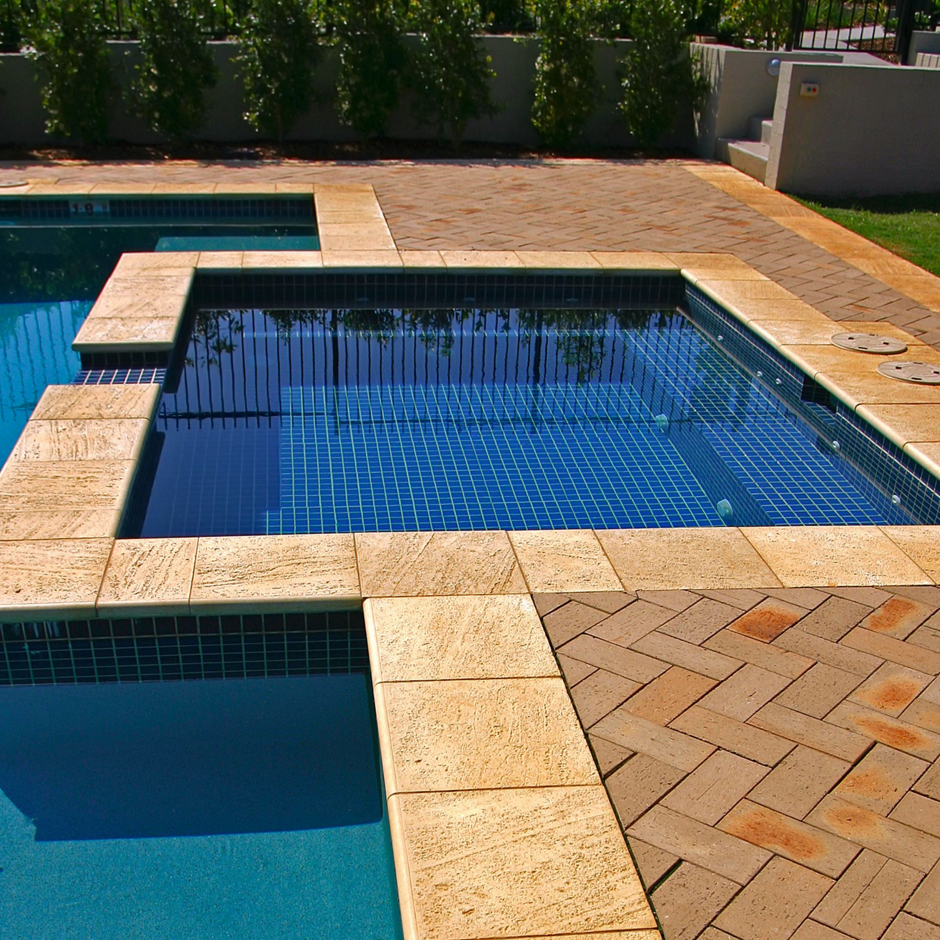 Piscine et spa en carrelage bleu, entourés de pavés et de briques de couleur claire, haies vertes en arrière-plan.
