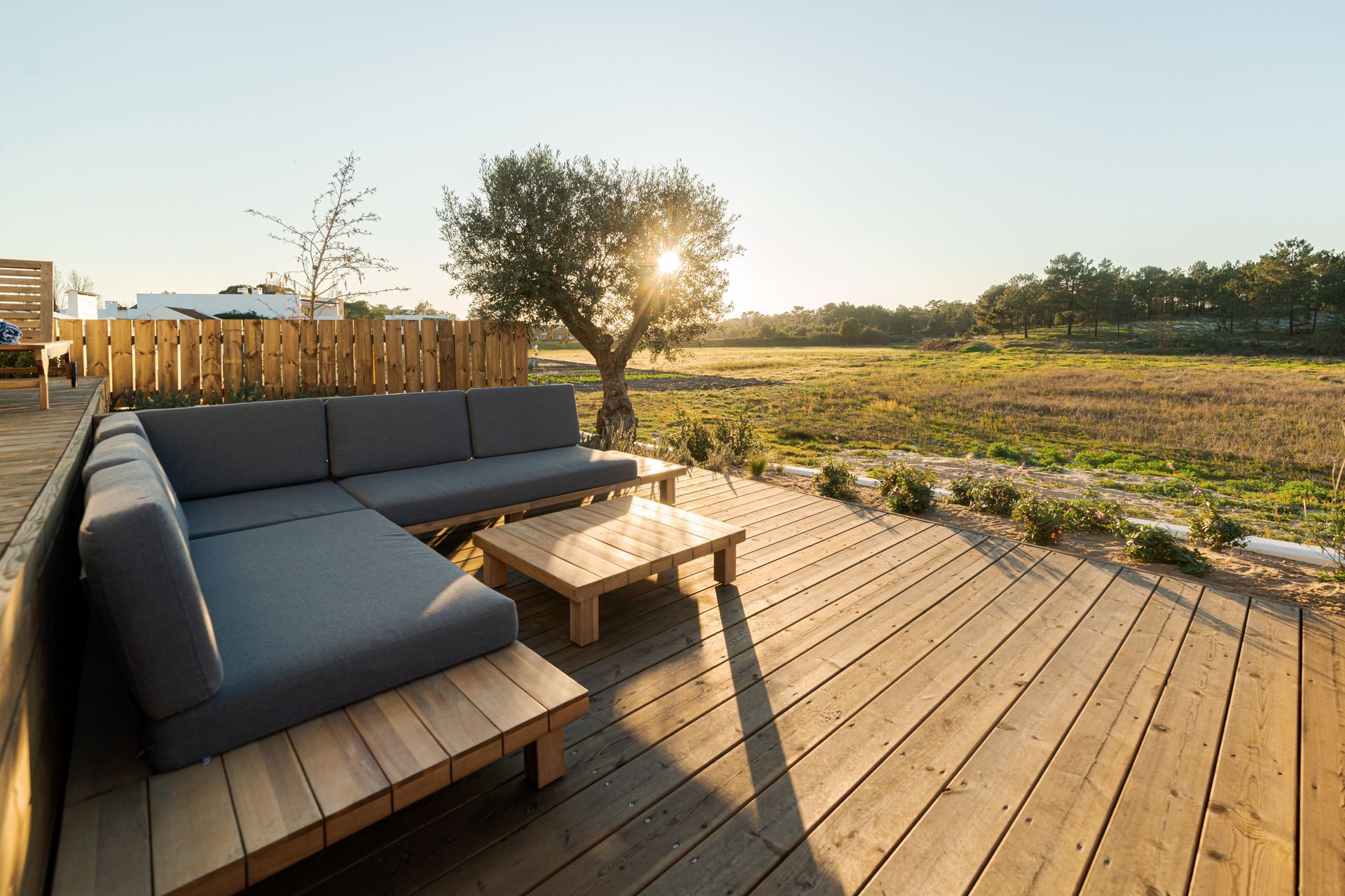 Terrasse en bois avec canapé gris et table donnant sur un champ, le soleil brille à travers un arbre.