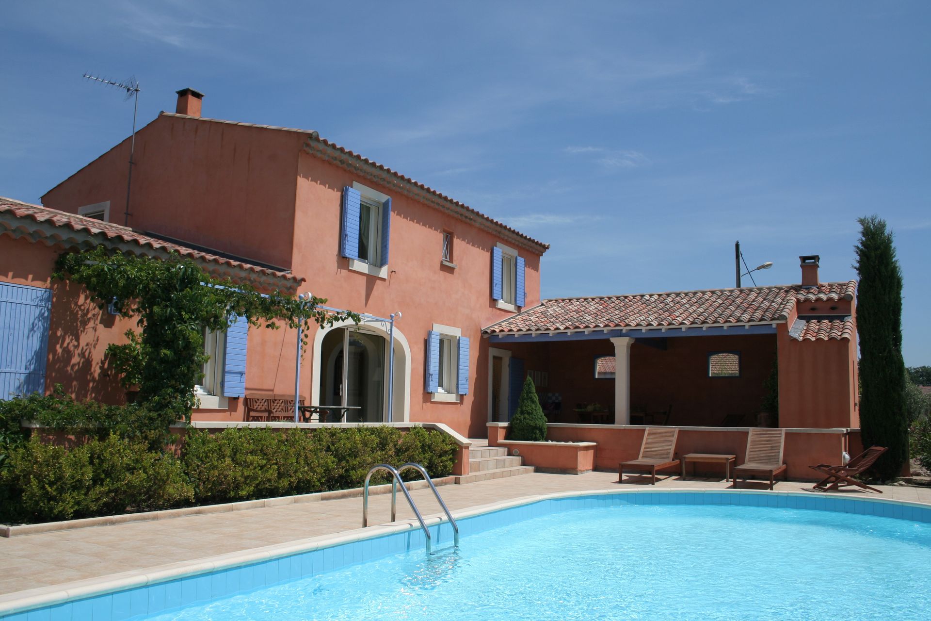 Maison en stuc rose avec volets bleus et piscine ; journée ensoleillée.
