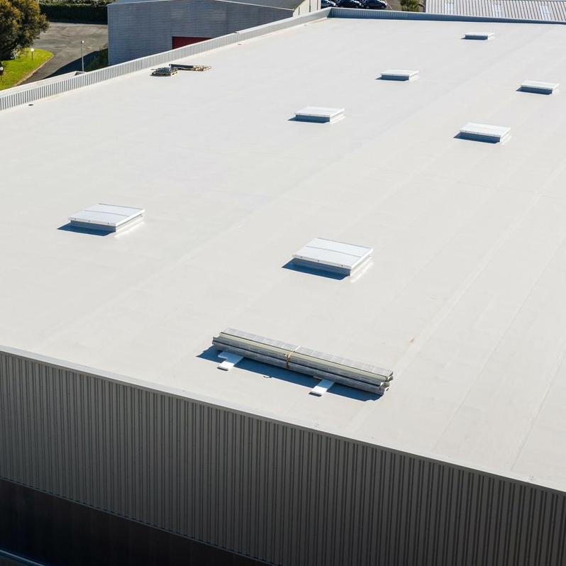Vue aérienne d'un grand bâtiment industriel de couleur claire avec un toit plat et plusieurs puits de lumière.