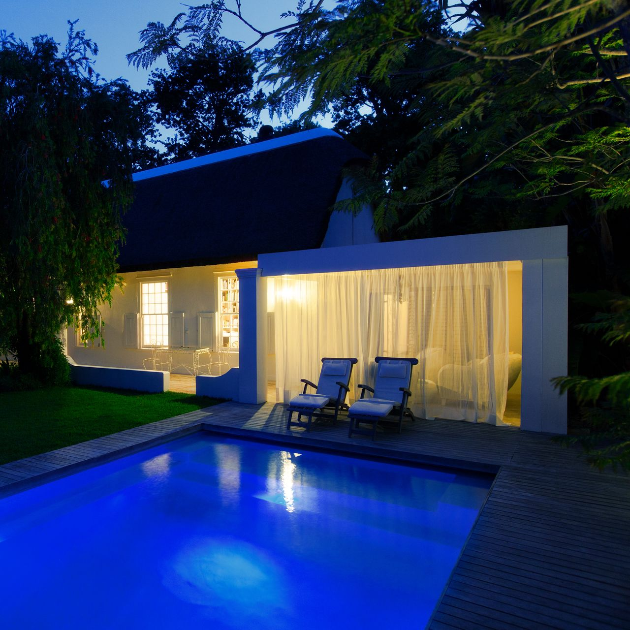 Maison et pool house au bord de la piscine au crépuscule avec chaises longues sous un éclairage doux.