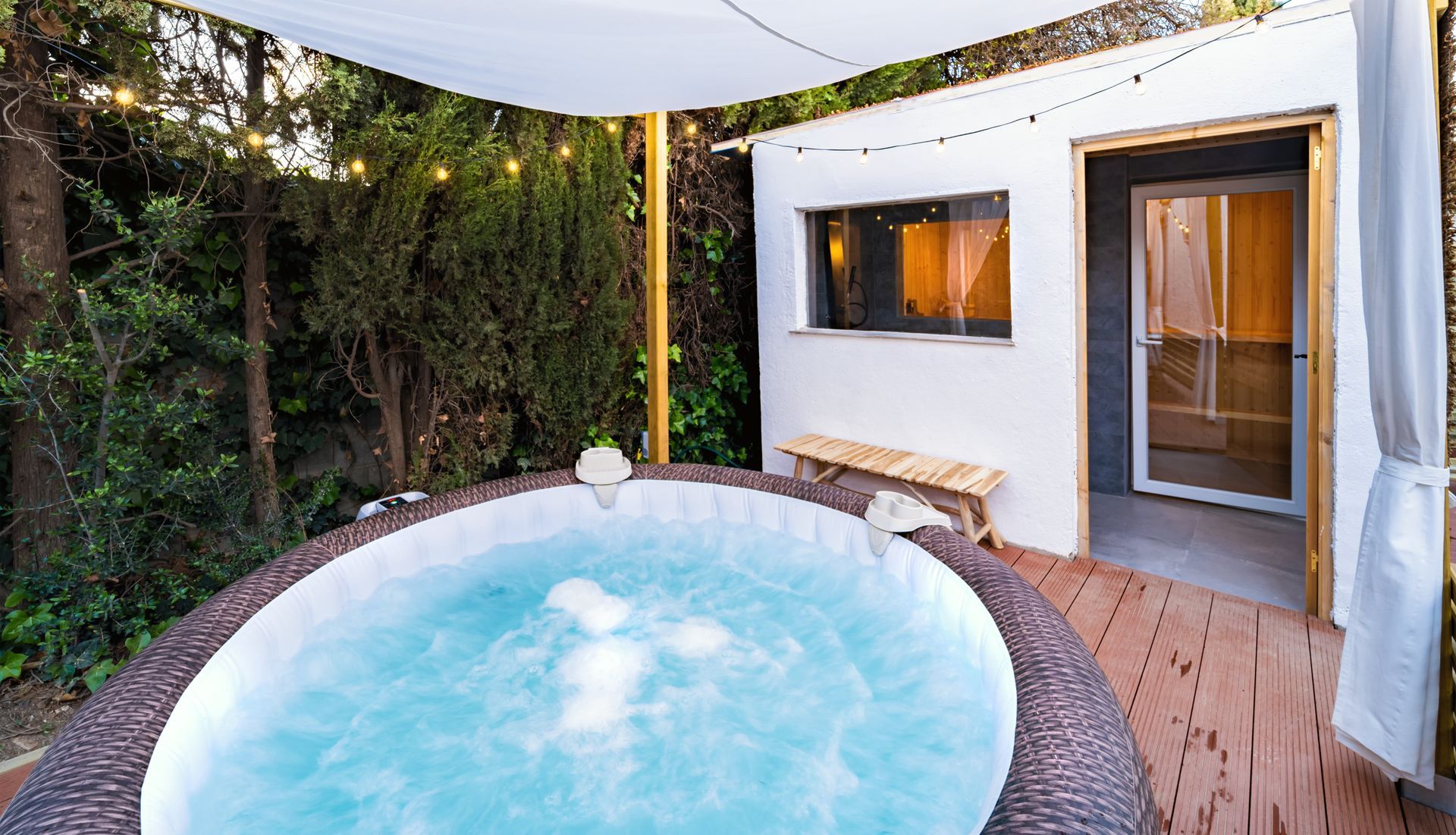 Jacuzzi sur une terrasse en bois à l'extérieur d'un bâtiment blanc, guirlandes lumineuses au-dessus.