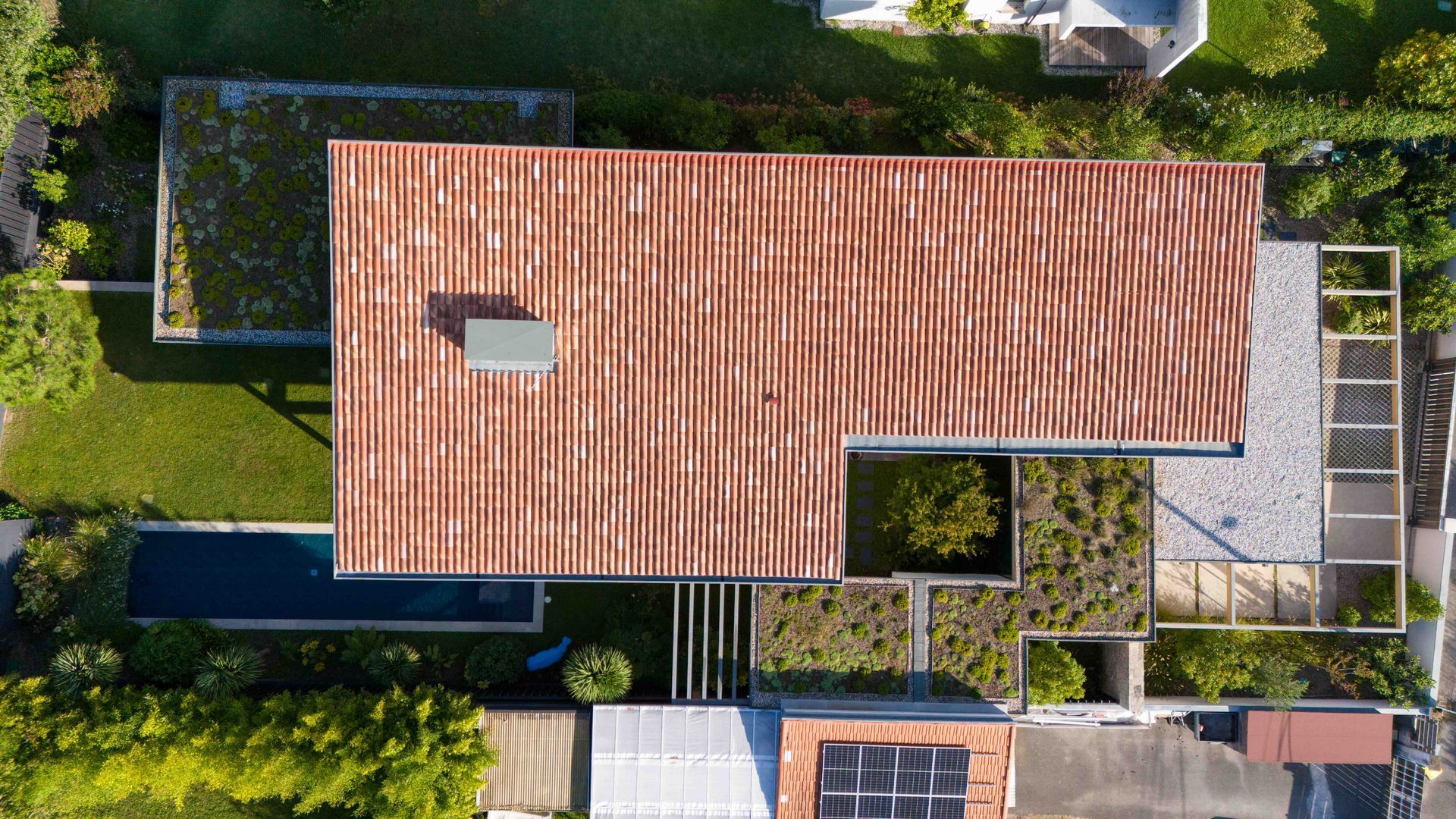 Vue aérienne d'une maison moderne avec un toit de tuiles rouges, entourée de toits verts, de jardins et d'une piscine.
