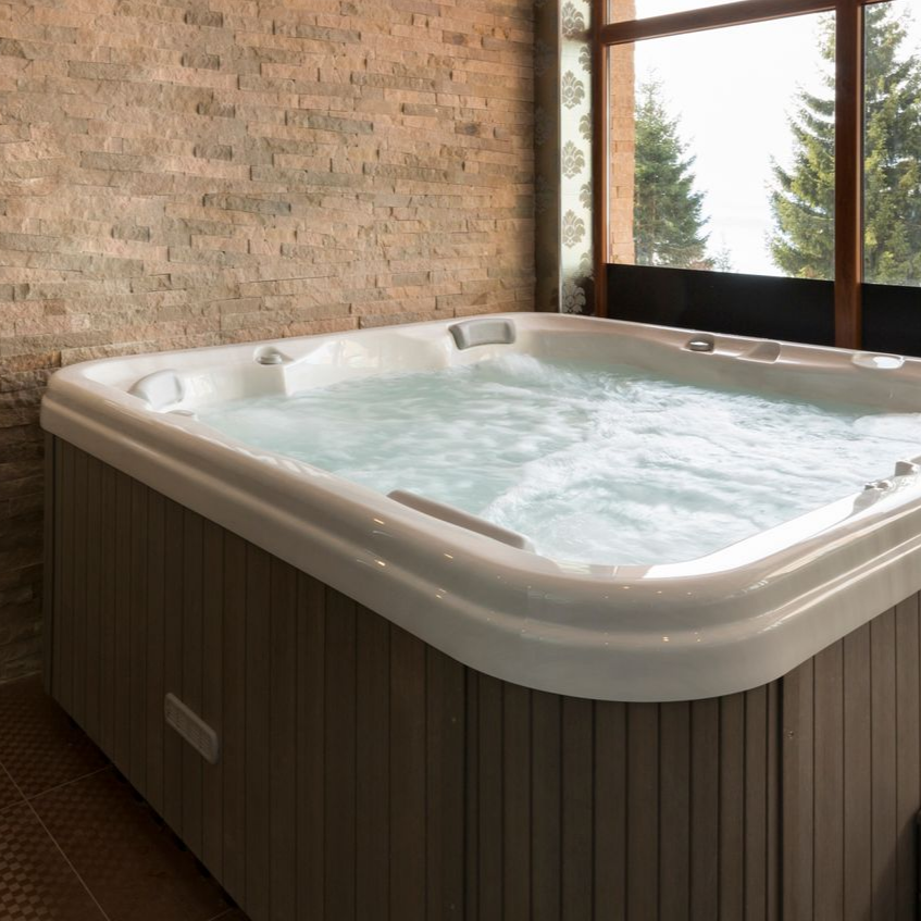 Bain à remous avec eau bouillonnante, placé contre un mur de pierre et une fenêtre avec des arbres en arrière-plan.