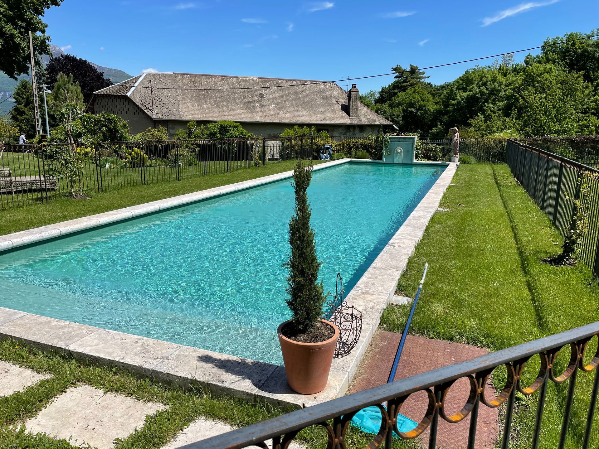 Longue piscine rectangulaire entourée d'herbe verte et d'un patio en pierre, avec une maison en arrière-plan.