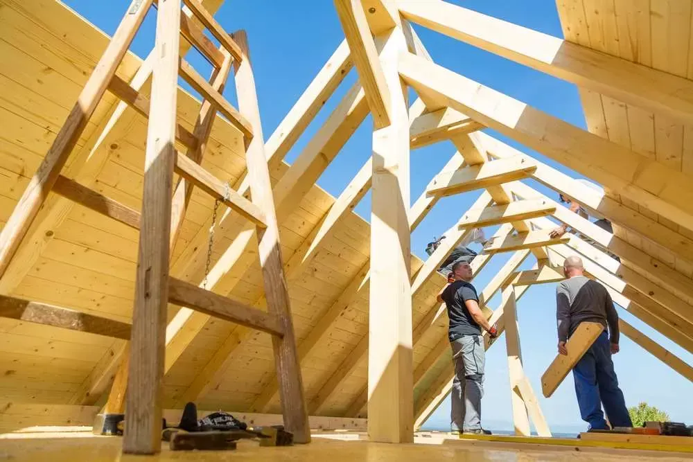 Des ouvriers du bâtiment assemblent des fermes de toit en bois contre un ciel bleu clair.