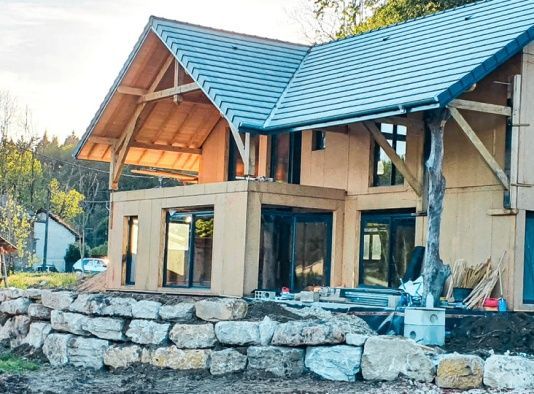 Maison neuve en bois en construction avec poutres apparentes, mur de soutènement en pierre et arbres environnants.