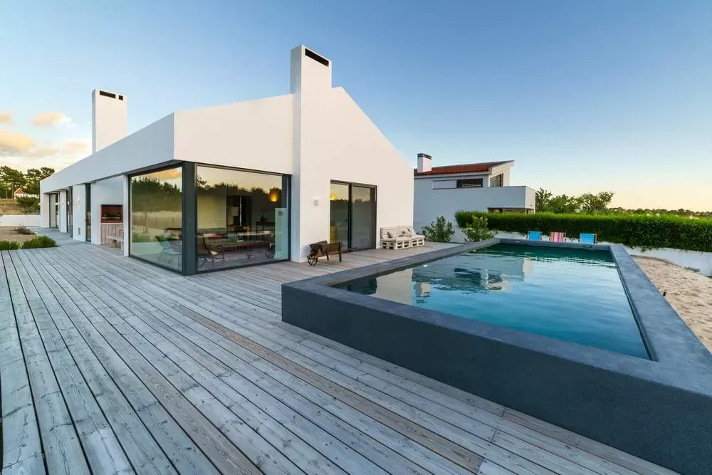 Maison blanche moderne avec piscine, terrasse en bois et ciel bleu clair.